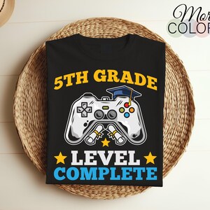 Pode incluir: Camiseta preta com um gráfico de um controlador de videogame e o texto "5th Grade Level Complete". O controlador tem um chapéu de formatura no topo.