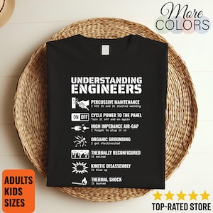 May include: Black t-shirt with white text and graphics. The shirt reads "UNDERSTANDING ENGINEERS" with humorous definitions of engineering terms. Includes "ADULTS KIDS SIZES" on a square orange label. The shirt is displayed on a woven basket.
