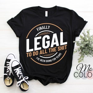May include: Black t-shirt with the text "FINALLY LEGAL TO DO ALL THE SHIT I'VE BEEN DOING FOR YEARS" in a circular design. The shirt is paired with black and white sneakers. The words "More COLORS" are also visible.