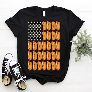 May include: Black t-shirt with a graphic of the American flag made of hot dogs. The hot dogs are yellow with red mustard and a white background.