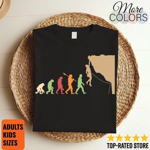 May include: Black t-shirt with a graphic showing human evolution, ending with a climber. The design includes the text "More Colors" and "ADULTS KIDS SIZES". The shirt is displayed on a woven basket.