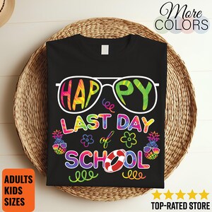 Può includere: T-shirt nero con una grafica colorata che recita "HAPPY LAST DAY OF SCHOOL". Il design include occhiali da sole, ananas, fiori e un salvagente. La maglietta è esposta su un cesto intrecciato. Il testo sulla maglietta è in stile tie-dye arcobaleno.