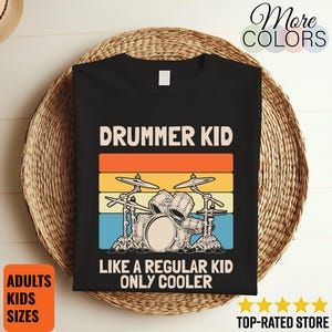 May include: Black t-shirt with a drum set graphic and the text "DRUMMER KID LIKE A REGULAR KID ONLY COOLER." The design includes orange, yellow, and blue stripes. The shirt is displayed on a woven basket.