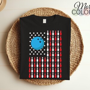 Może przedstawiać: Czarny T-shirt z piłką do kręgli i kręglami ułożonymi w kształt amerykańskiej flagi. Piłka do kręgli jest niebieska, a kręgle są czerwone i białe.