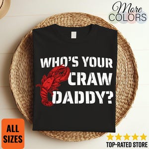 以下が含まれることがあります： 「WHO'S YOUR CRAW DADDY？」の白い文字と赤いザリガニのイラストがプリントされた黒いTシャツ。Tシャツは編みかごの上に置かれています。「ALL SIZES」と「TOP-RATED STORE」の文字も含まれています。