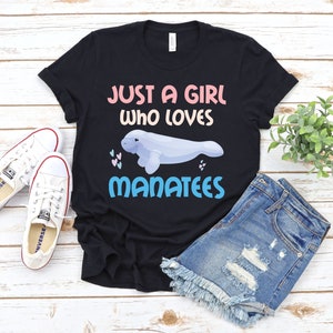 Puede incluir: Camiseta negra con el texto "Just a girl who loves manatees" en rosa, azul y blanco. Un dibujo animado de manatí se representa junto al texto.