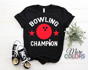 May include: Black t-shirt with the words "BOWLING CHAMPION" in white, featuring a red bowling ball and two red stars. A pair of black and white sneakers are in the foreground. The shirt design includes a bowling pin.