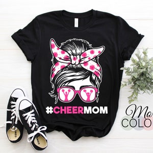 Può includere: T-shirt nero con grafica di una donna con fascia floreale rosa e occhiali con un design da cheerleader. Il testo "#CHEERMOM" è sotto la grafica. Scarpe da ginnastica nere e bianche in primo piano.