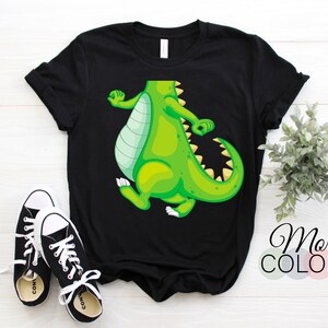 Op de afbeelding: Zwart T-shirt met een cartoon dinosaurus in groen en geel. De dinosaurus staat in een speelse pose. De tekst "More Colors" is zichtbaar. Een paar zwart-witte sneakers staan op de voorgrond.
