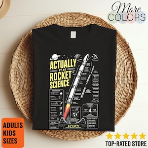 Peut inclure: Un t-shirt noir avec un motif blanc et jaune représentant une fusée et le texte "ACTUALLY IT IS ROCKET SCIENCE". Le design comprend des schémas techniques, des équations scientifiques et des étiquettes détaillant les composants de la fusée.