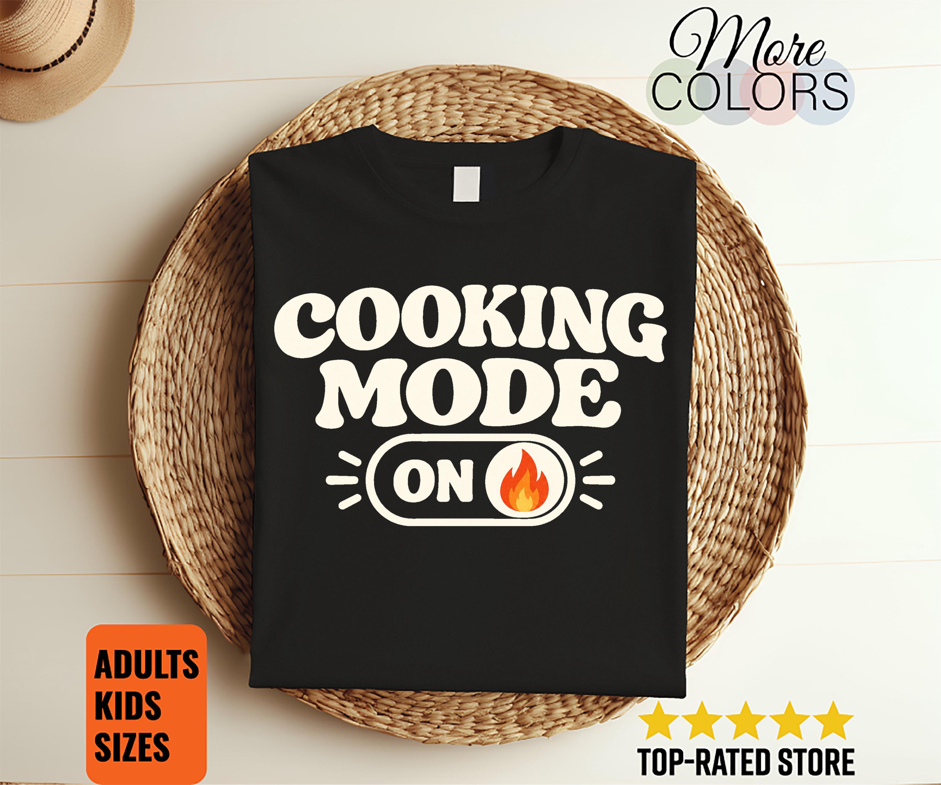 以下が含まれることがあります： 黒いTシャツで、白い文字で「COOKING MODE」と書かれ、その下に「ON」という文字と炎のアイコンが描かれたトグルスイッチのグラフィックがあります。デザインはシャツの胸の中央に配置されており、シンプルなクルーネックと半袖が特徴です。