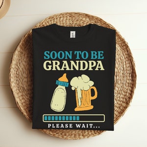 May include: Black t-shirt with the text "Soon to be Grandpa" in blue and yellow.  A baby bottle and a beer mug are pictured below the text. A progress bar with the text "Please wait..." is at the bottom of the shirt.