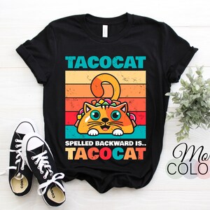 May include: Black t-shirt with a retro-style graphic of a cat holding a taco. The text "Tacocat" is above the cat and the text "Spelled backward is... Tacocat" is below the cat.