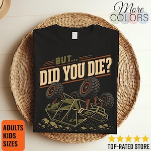 May include: A black t-shirt featuring a graphic of an overturned off-road vehicle with large tyres and suspension details. Above the vehicle, the text reads BUT... DID YOU DIE? in a bold, retro-style font with olive green and cream-coloured accents.
