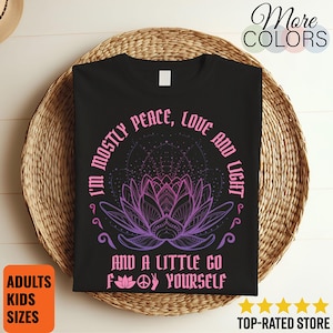 May include: Black t-shirt featuring a purple and pink lotus flower design. The text on the shirt says "I'm mostly peace, love and light, and a little go f*** yourself." The shirt is displayed on a woven basket.