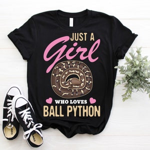 Op de afbeelding: Zwart T-shirt met de tekst "Just a Girl Who Loves Ball Python" in roze en beige. Het ontwerp bevat een afbeelding van een opgerolde koningspython. Een paar zwart-witte sneakers staan op de voorgrond.