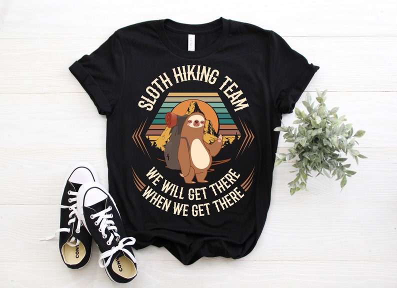 May include: Black t-shirt with a retro-style graphic of a sloth wearing a backpack and hiking in the mountains. The text reads "Sloth Hiking Team We Will Get There When We Get There".