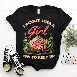 May include: Black t-shirt featuring a graphic of a tent, trees, and mountains. The text "I scout like a girl try to keep up" is printed on the shirt.
