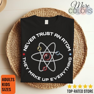 Peut inclure: T-shirt noir avec un motif d'atome blanc et le texte "NEVER TRUST AN ATOM THEY MAKE UP EVERYTHING". Le t-shirt est présenté sur un panier en osier. Le texte supplémentaire comprend "More COLORS", "ADULTS KIDS SIZES" et "TOP-RATED STORE".