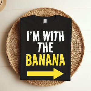 Puede incluir: Camiseta negra con texto blanco que dice "Estoy con el plátano" y una flecha amarilla apuntando hacia la derecha.
