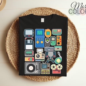 Puede incluir: Una camiseta negra con un gráfico colorido de dispositivos electrónicos vintage, que incluye un televisor, una consola de videojuegos, un reproductor de casetes, una cámara, un teléfono y un tocadiscos.