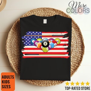 May include: Black t-shirt featuring a distressed American flag design with pool balls and cues. The number 8 ball is prominently displayed. The shirt also has the text "More COLORS", "ADULTS KIDS SIZES", and "TOP-RATED STORE".