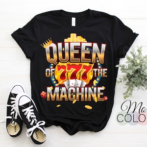 May include: Black t-shirt with a graphic that reads "QUEEN OF THE MACHINE" in gold and red. The design includes a crown, playing cards, dice, and gold coins. A pair of black and white sneakers are in the foreground.