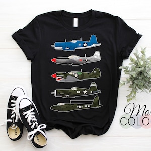 以下が含まれることがあります： 黒いTシャツ。5機のヴィンテージ飛行機のイラストが描かれており、青、シルバー、緑、オリーブグリーンなど様々な色があります。「More Colors」の文字がシャツの右側に表示されています。