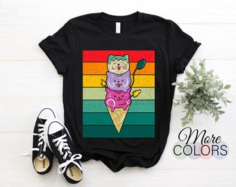 May include: Black t-shirt featuring a colorful graphic of three cat-shaped ice cream scoops on a cone, set against a striped background of red, orange, yellow, green, and blue. The design includes a spoon and smiling faces. The words "More Colors" are also visible.