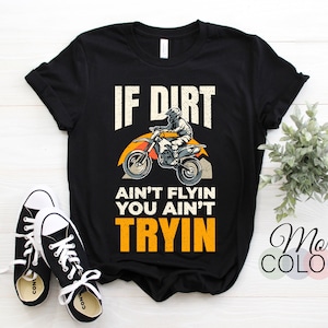 Peut inclure: T-shirt noir avec l'inscription "IF DIRT AIN'T FLYIN YOU AIN'T TRYIN" et un motif de moto tout-terrain. Le design comprend des accents orange, jaune et blanc. Une paire de baskets noires et blanches est au premier plan.