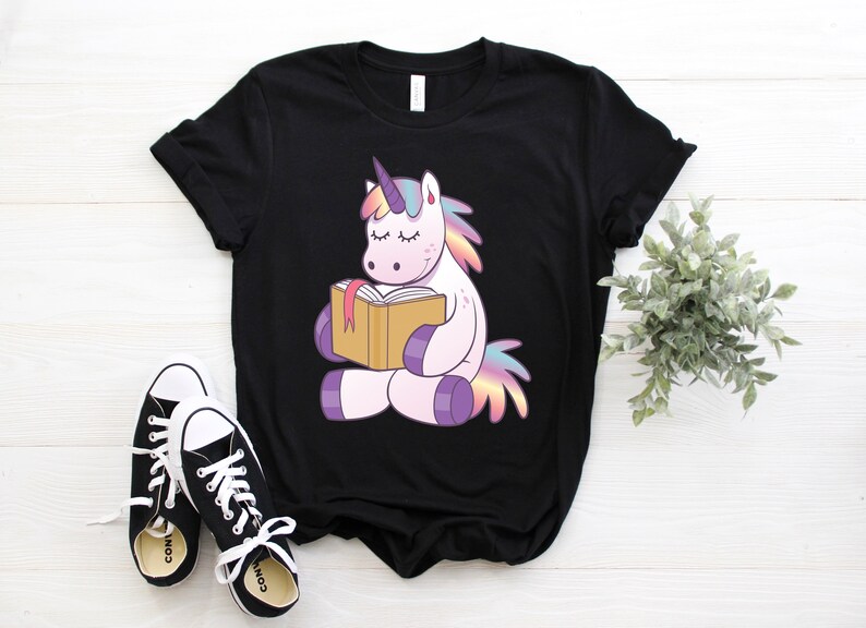 Pode incluir: Camiseta preta com um unic&oacute;rnio de desenho animado lendo um livro. O unic&oacute;rnio tem uma crina e cauda coloridas, com um chifre roxo. T&ecirc;nis preto e branco em primeiro plano e uma pequena planta &agrave; direita.