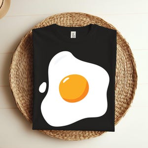 Pode incluir: Camiseta preta com um gráfico branco de um ovo frito. A gema é amarela.