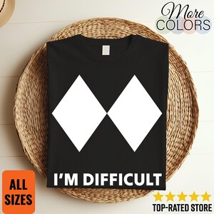 May include: A black t-shirt featuring two large white diamond shapes positioned side by side above the white text that reads I'M DIFFICULT. The shirt has a crew neckline and short sleeves, presenting a bold graphic design on the front center.