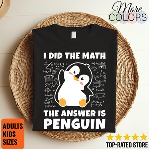 May include: Black t-shirt featuring the text "I DID THE MATH THE ANSWER IS PENGUIN" and a cartoon penguin. The penguin is black and white with orange feet and a yellow beak. The shirt is displayed on a woven basket.