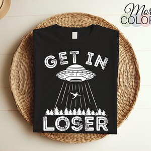 Puede incluir: Camiseta negra con un gráfico blanco de un OVNI con el texto "GET IN LOSER" debajo.