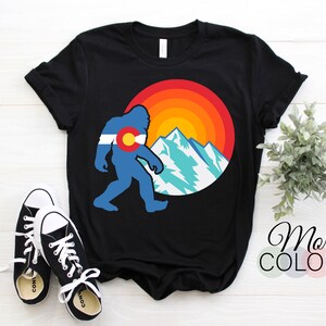 May include: Black t-shirt with a graphic of a Bigfoot walking in front of a mountain range with a sunset behind it. The Bigfoot is wearing the Colorado flag on its chest.