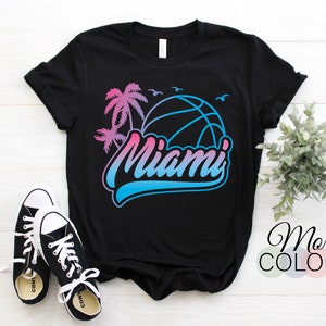 Puede incluir: Camiseta negra con un diseño degradado en rosa y azul que presenta una pelota de baloncesto con palmeras y la palabra "Miami".