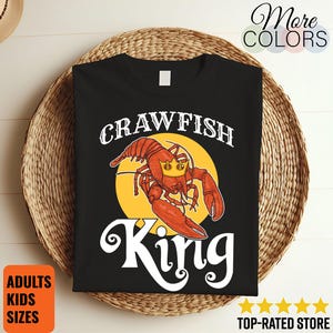 May include: Black t-shirt featuring a red crawfish wearing a crown, set against a yellow circle. The text "CRAWFISH KING" is displayed in white, with additional text indicating adult and kids sizes. The shirt is on a woven surface.