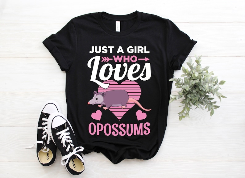 Puede incluir: Camiseta negra con el texto "JUST A GIRL WHO LOVES OPOSSUMS". El dise&ntilde;o presenta un coraz&oacute;n rosa con un fondo a rayas, una zarig&uuml;eya de dibujos animados y corazones rosas. Zapatillas negras y blancas en primer plano.