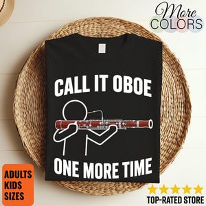 May include: A black t-shirt featuring a white stick figure illustration holding a bassoon. The text above the figure reads CALL IT OBOE and the text below reads ONE MORE TIME in bold white capital letters. The design is centred on the shirt.