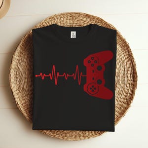 Puede incluir: Camiseta negra con un gráfico rojo de un mando de videojuego y una línea de latido del corazón. El texto "I WAS" es visible en la parte posterior de la camiseta.