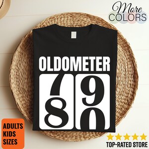 May include: Black t-shirt with the word "OLDOMETER" above the numbers 79 and 80 in white. The shirt is displayed on a woven basket. Additional text includes "More Colors", "Adults Kids Sizes", and "Top-Rated Store".