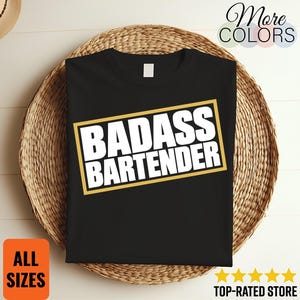 Op de afbeelding: Een zwart t-shirt met korte mouwen en een rechthoekige afbeelding met een gouden rand en de witte tekst "BADASS BARTENDER" in het midden op de borst. Het shirt heeft een standaard ronde hals en een effen zwarte achterkant.