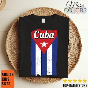 Może przedstawiać: Czarna koszulka z postarzanym wzorem flagi Kuby i napisem "Cuba". Flaga przedstawia czerwony trójkąt z białą gwiazdą oraz niebiesko-białe paski. Koszulka jest prezentowana na wiklinowym koszu.
