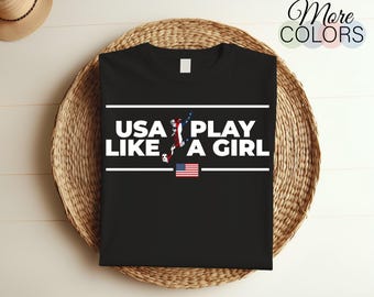 May include: Black t-shirt with white text that reads "USA Play Like A Girl" and a graphic of a soccer player. The shirt also features a small American flag.