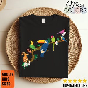 Peut inclure: T-shirt noir avec une illustration colorée d'oiseaux sur une branche, dont un toucan et un perroquet. Le motif comprend des fleurs blanches et le texte "More Colors", "Adults Kids Sizes", et "Top-Rated Store".