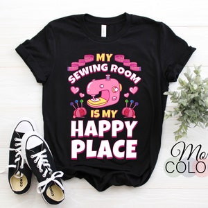 Peut inclure: T-shirt noir avec l'inscription "MY SEWING ROOM IS MY HAPPY PLACE" et un motif de machine à coudre rose. Le motif comprend des cœurs roses, un mètre ruban et des coussins à épingles. Des baskets noires et blanches sont au premier plan.