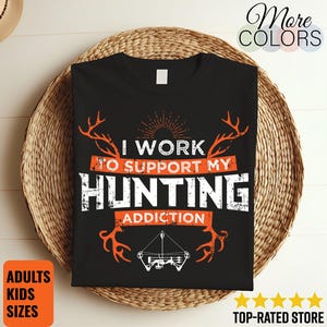 Puede incluir: Camiseta negra con el texto "I WORK TO SUPPORT MY HUNTING ADDICTION" en blanco y naranja. El diseño incluye gráficos de cuernos y un arco y flecha. La camiseta se muestra en una cesta tejida.