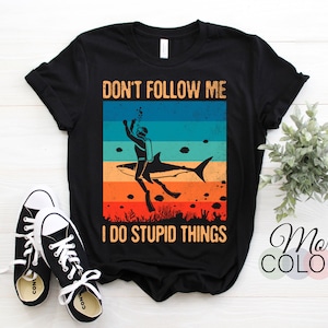 Pode incluir: Camiseta preta com um gráfico de estilo vintage de um mergulhador montando um tubarão. O texto "Não me siga, eu faço coisas estúpidas" está impresso na camiseta.