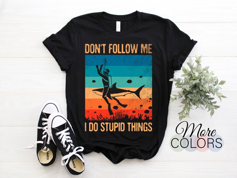 Może przedstawiać: Czarny T-shirt z grafiką w stylu vintage przedstawiającą nurka jadącego na rekinie. Na koszulce nadrukowany jest tekst "Nie podążaj za mną, robię głupie rzeczy".